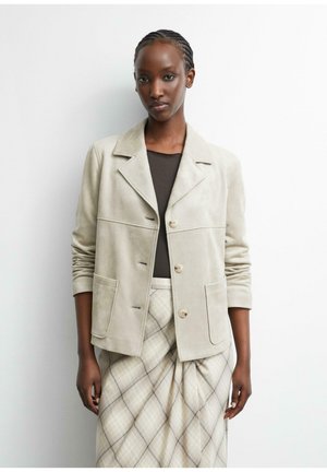 Frau mit geflochtenem Haar, die eine beige Jacke über einem schwarzen Oberteil und einem beige karierten Rock trägt, steht vor einem weißen Hintergrund.