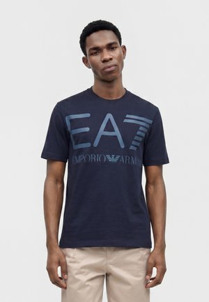 Jeune homme portant un t-shirt bleu marine Emporio Armani EA7 et un pantalon beige, debout devant un fond clair uni.
