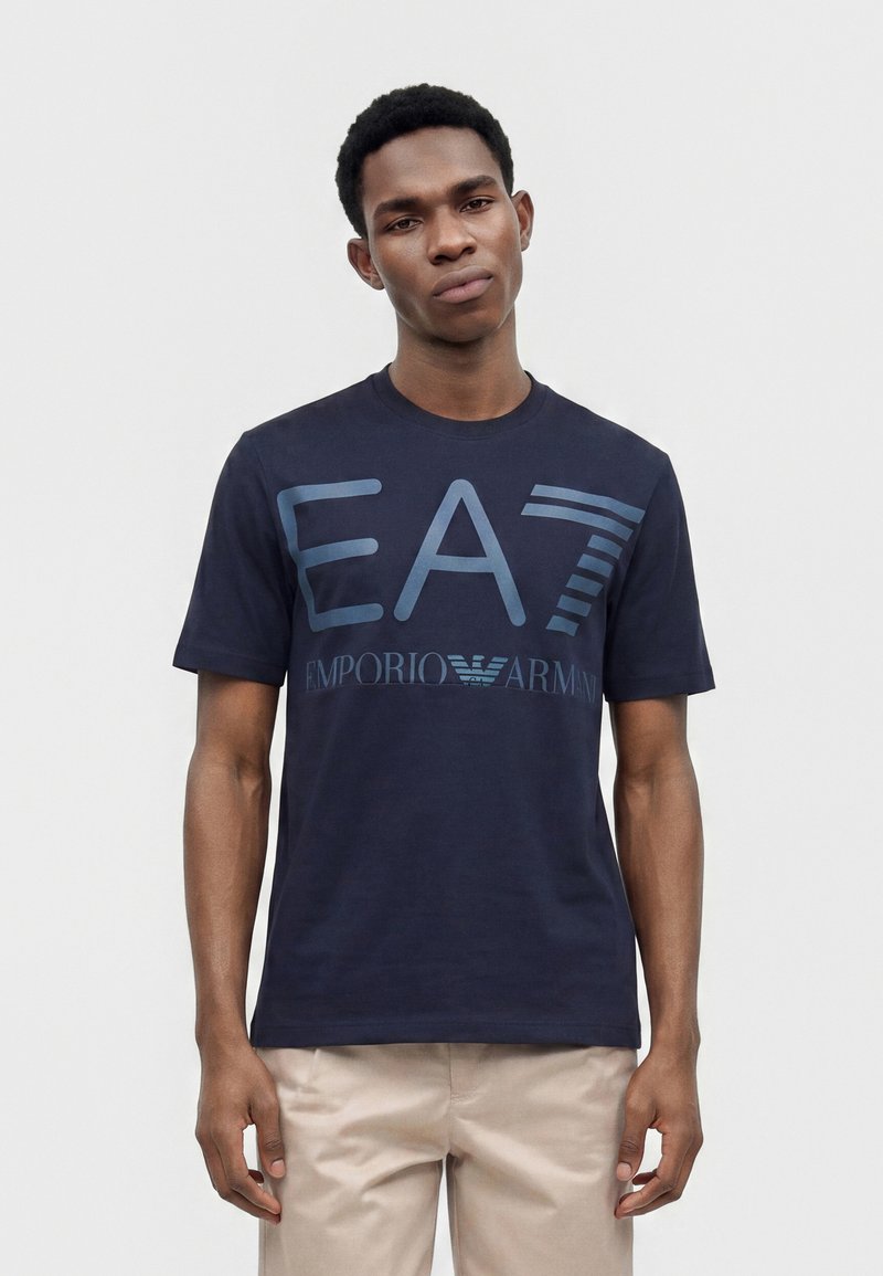Νεαρός άνδρας που φοράει μπλούζα Emporio Armani EA7 σε ναυτικό μπλε και μπεζ παντελόνι, στέκεται μπροστά σε απλό ανοιχτόχρωμο φόντο.