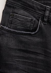 Jeans de mezclilla negra con un bolsillo frontal con un borde deshilachado, un ligero desvanecimiento y detalles de costura prominentes a lo largo de la cinturilla y el bolsillo.