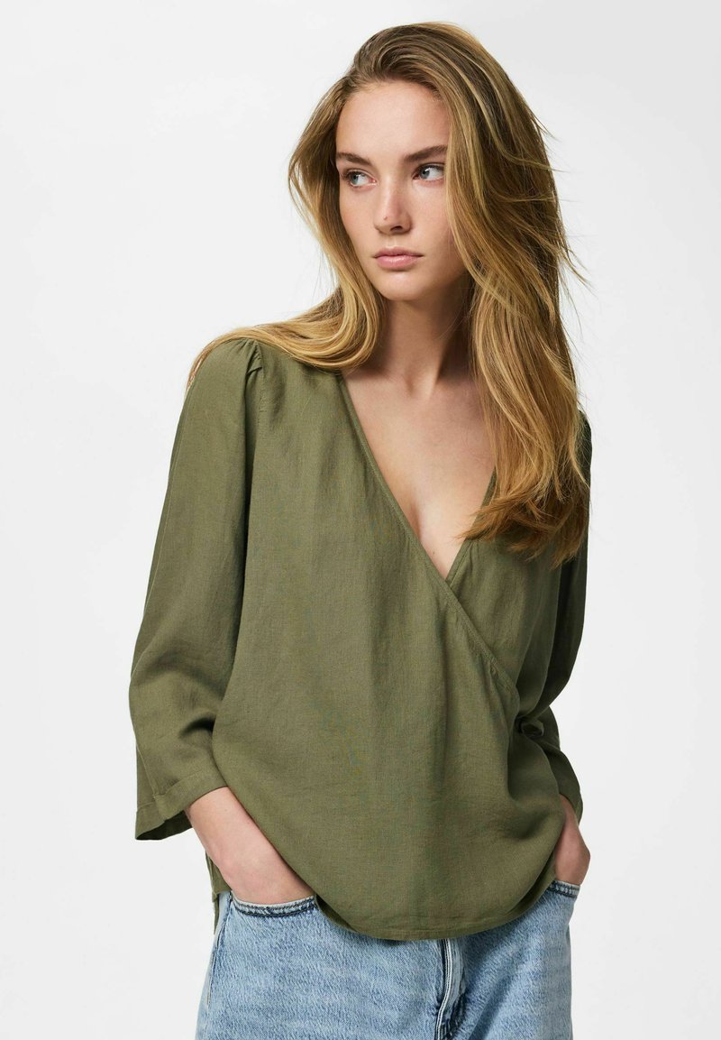 Jeune femme aux longs cheveux blonds portant un chemisier vert olive à col en V et un jean bleu clair, les mains dans les poches, regardant sur le côté.