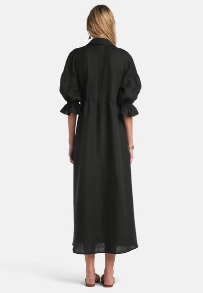 Woman standing with back visible, wearing a black midi dress with puffed elbow-length sleeves and sandals on a white background.