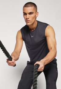 Tricou sportiv fără mâneci de culoare gri închis, fabricat dintr-un material neted, cu un logo pe piept. Modelul ține o frânghie groasă de culoare neagră.