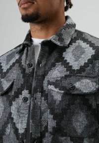 Man draagt een grijs-zwart wollen patroon jack met knopen over een wit shirt, waarbij de kraag en borst zichtbaar zijn.