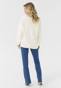 Cremearmiger Pullover mit lockerer Passform und gerippten Bündchen, kombiniert mit figurbetonten blauen Jeans. Das Modell hat welliges Haar und ist von hinten zu sehen.