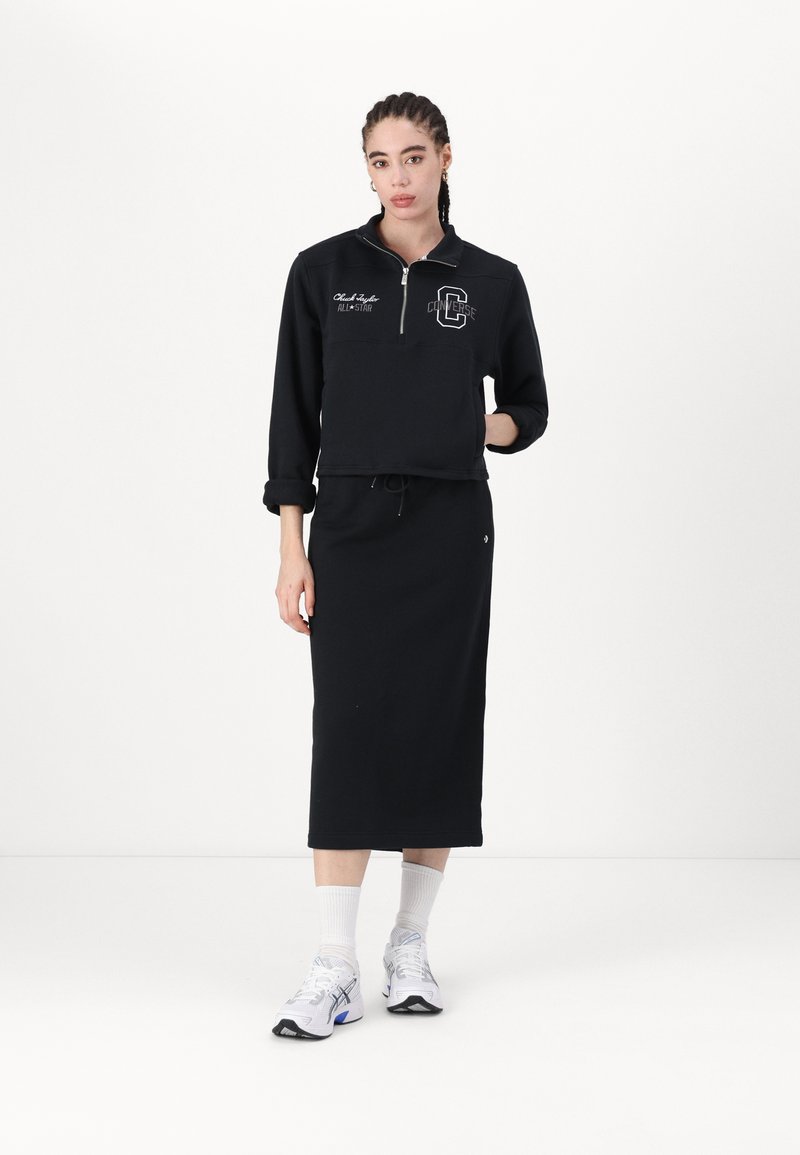 Sweatshirt noir avec col zippé mi-hauteur, logo brodé et jupe longue noire. Porté avec des chaussures de sport blanches et des chaussettes légères. Design minimaliste.