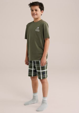 Olijfgroene katoenen T-shirt met korte mouwen, gecombineerd met groene geruite shorts met witte en marineblauwe accenten, en grijze geribbelde sokken.