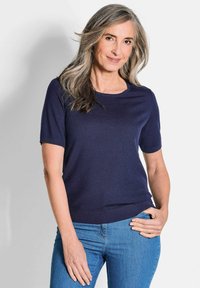Marineblaue kurzärmlige Strickjacke aus weichem Material; mit Rundhalsausschnitt und gerafftem Schnitt, kombiniert mit hellblauen Jeans.