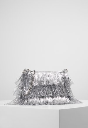 Sac à main à franges métalliques argentées avec une bandoulière chaîne, présenté sur un socle blanc devant un fond gris uni.