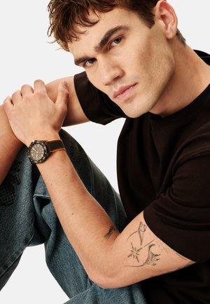 Ung mand med tatoveringer iført sort skjorte og jeans, hviler armen på knæet og viser sit smartwatch med sort og orange rem.
