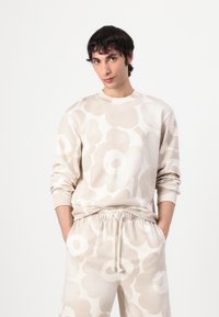 Beige Sweatshirt mit großen hellen Blumenmustern, geripptem Rundhalsausschnitt und langen Ärmeln. Kombiniert mit passenden Jogginghosen mit Kordelzug.