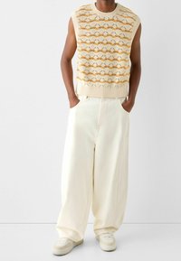 Stickad ärmlös topp i krämfärg med gult vågmönster; lösa vita byxor; neutrala sneakers; avslappnad passform, minimalistisk design.