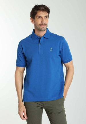 Homme portant un polo bleu à manches courtes avec un petit logo sur la poitrine et un pantalon vert olive, debout avec une main dans la poche.