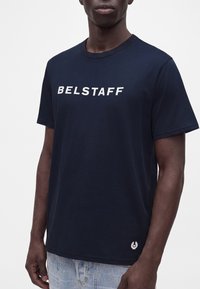 Άνδρας που φοράει μπλε μπλουζάκι Belstaff με λευκό λογότυπο στο στήθος και ανοιχτό μπλε τζιν, φωτογραφημένος από τον λαιμό μέχρι τους γοφούς.