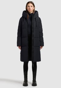 Sort puffetjakke med et langt, lige design, der har hætte, frontlynlås og justerbare snore. Glat tekstur, quiltede detaljer.