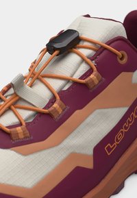 Detailní záběr jazyka tenisky s oranžovými elastickými tkaničkami a černým jezdcem na béžové a vínové látce s nápisem "LOWA" na straně.