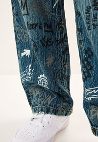 Jeans di denim blu decorati con vari motivi in stile graffiti in nero, grigio e bianco. Abbinati a sneakers bianche per creare contrasto.