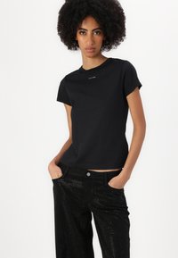 Crna pamučna majica s kratkim rukavima i okruglim izrezom; na prsima sadrži bijeli "Calvin Klein" logo. Uparena s crnim perforiranim hlačama.