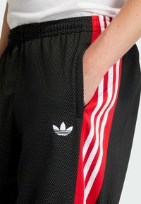 Svarta träningsbyxor med en nätliknande textur, utrustade med röda sidoremmar och vit Adidas-logotyp. Resårlinning och sidofickor ingår.