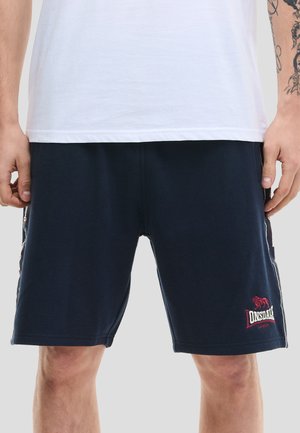 Uomo che indossa pantaloncini blu navy Lonsdale London e una maglietta bianca semplice, mostrando il torso inferiore e la parte superiore delle gambe con il braccio destro tatuato.