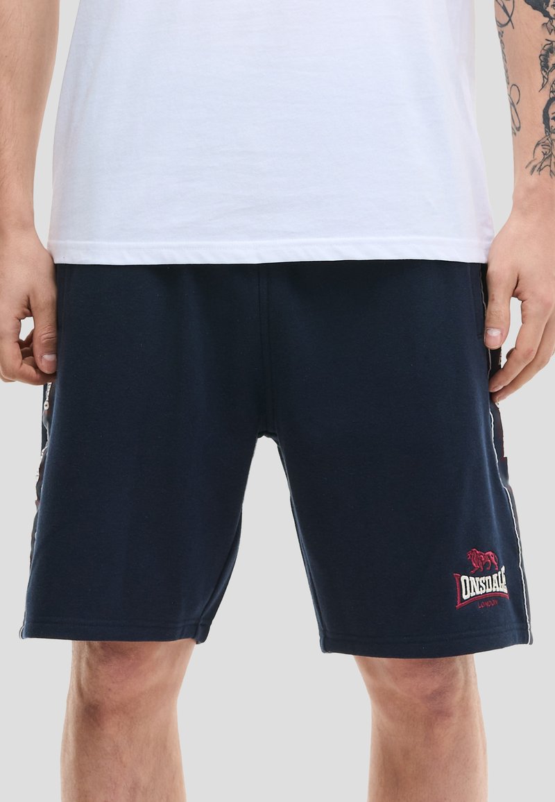 Mężczyzna w granatowych szortach Lonsdale London i białej, gładkiej koszulce, ukazujący dolną część tułowia i górną część nóg, z wytatuowaną prawą ręką.