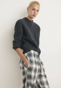 Mørk grå kabelstriktrøje med høj krave, parret med grå og hvid ternede bukser, der har sidelommer og en løs pasform.