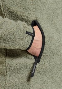 Fleecetröja i olivgrön färg med en strukturerad yta. Har en svart dragkedjeficka på ärmen med en märkesetikett.