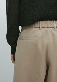 Pantaloni beige in tessuto liscio, con una tasca posteriore caratterizzata da una cucitura discreta. Abbinati a un maglione scuro e strutturato.