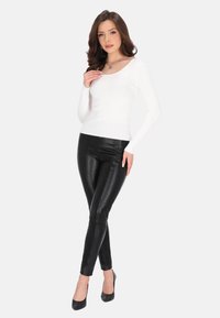 Top alb cu dungi, cu mânecă lungă, asortat cu pantaloni strâmți negri din piele ecologică și tocuri negre înalte. Modelul stă cu o mână aproape de guler.