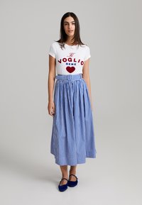Weißes T-Shirt mit rotem und blauem grafischem Text, kombiniert mit einem knielangen Rock aus blau-weißem Vichy-Karo. Blaue Ballerinas runden das Outfit ab.