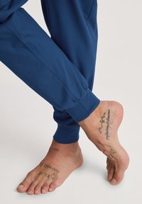 Pantalons de jogging bleu marine en tissu doux, avec des poignets élastiques, portés pieds nus avec des tatouages visibles sur la peau.