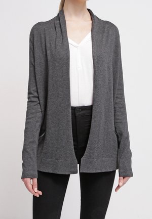 Cardigan grigio aperto davanti in tessuto morbido con una vestibilità rilassata, caratterizzato da maniche lunghe e due tasche frontali. Indossato sopra una maglietta bianca e pantaloni neri.