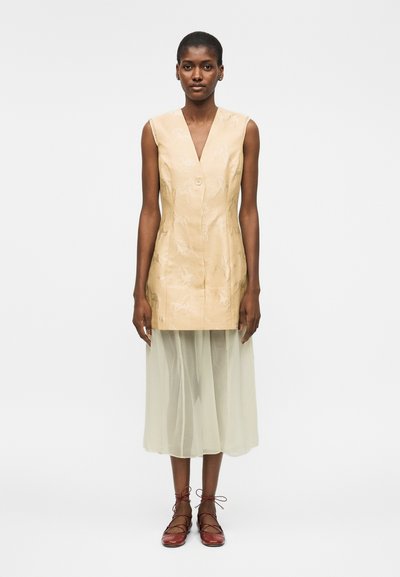 Mujer que lleva un chaleco beige sin mangas abotonado sobre una falda midi blanca translúcida y zapatos planos granate con cordones, de pie frente a un fondo liso.