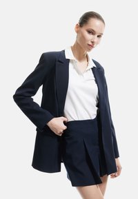 Marineblå blazer med skræddersyet design og klippekrave, parret med en hvid polo-shirt og en matchende marineblå plisseret nederdel.