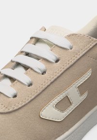 Μπεζ σουέτ αθλητικό παπούτσι με λευκά κορδόνια και ραμμένο λευκό λογότυπο στο πλάι, με λεπτομερή κοντινή λήψη του πάνω μέρους.