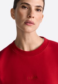 Piros crewneck pulóver, texturált felülettel, kontrasztos cérnával hímzett "RCHRYL" logóval. Közeli nézet a szövet és a nyakkivágás részleteiről.