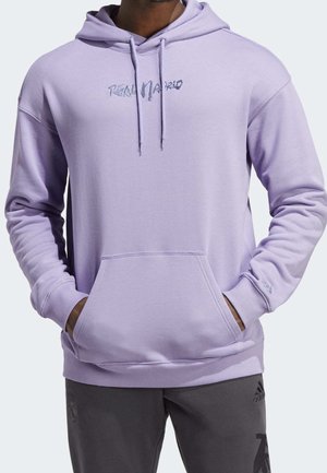 Homme portant un sweat à capuche violet clair avec le texte "Real Madrid" et un pantalon Adidas noir, mains dans la poche du sweat, debout devant un fond uni.