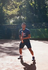 Τενίστας με μπλε μπλούζα και μαύρο σορτς κρατάει μια ρακέτα σε ένα κοκκινόχωμα γήπεδο, περιτριγυρισμένος από πράσινο και ηλιοφάνεια.