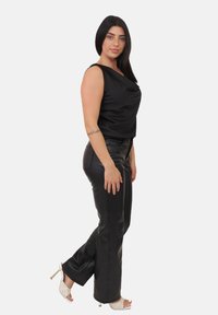 Donna con capelli lunghi e scuri, indossando un top senza maniche nero e pantaloni in pelle nera, posa di lato con sandali con tacco alto bianchi su uno sfondo bianco.
