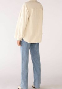 Camicia in denim color crema a maniche lunghe con orlo curvo e polsini con bottone, abbinata a jeans azzurri chiari e scarpe color crema.
