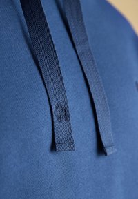 Nahaufnahme von blauem Stoff mit zwei dunkelblauen, gewebten Kapuzenkordeln, von denen eine ein kleines aufgesticktes Logo nahe dem Ende zeigt.