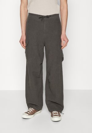 Person, der dunkelgraue, weit geschnittene Cargohosen mit Fronttaschen und braune Low-Top-Sneaker trägt, steht vor einem weißen Hintergrund.