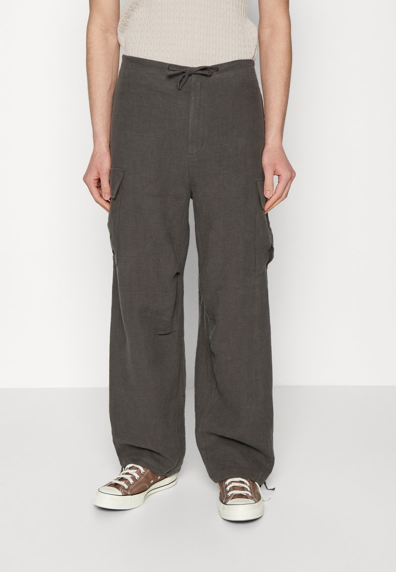 Personne portant un pantalon cargo gris foncé à jambes larges avec des poches avant et des baskets basses marron, debout devant un fond blanc.