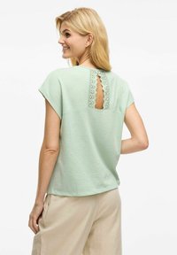 Krótki rękaw miętowa bluzka z koronkowym wycięciem z tyłu, charakteryzująca się luźnym krojem i miękką fakturą. Lekki, przewiewny materiał.