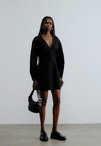 Μαύρο φόρεμα με μακριά μανίκια, βαθύ ντεκολτέ σε σχήμα V και φουσκωτή στεφάνη. Συνδυασμένο με μαύρη τσάντα και μαύρα παπούτσια. Απλός, μινιμαλιστικός σχεδιασμός.