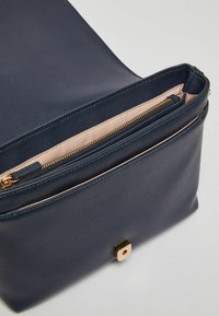 Borsa a mano in pelle blu navy con zip dorata, foderata in rosa, e un dettaglio in metallo dorato a forma quadrata sul davanti. Design strutturato, apertura superiore.