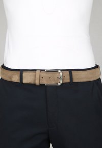 Lichtbruine suède riem met een zilveren gesp, gedragen over een marineblauwe broek en een witte ingestopte blouse op de taille van een persoon.