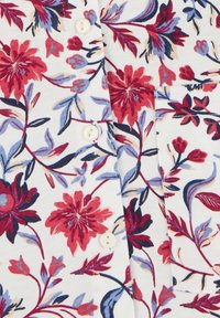 Tessuto bianco con un motivo floreale rosso e viola, con bottoni bianchi e una tasca frontale cucita sulla camicia.