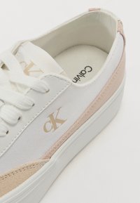 Baskets blanches Calvin Klein avec des accents beiges, en tissu et en daim, bout rond, semelle plate et détail de logo sur le côté.