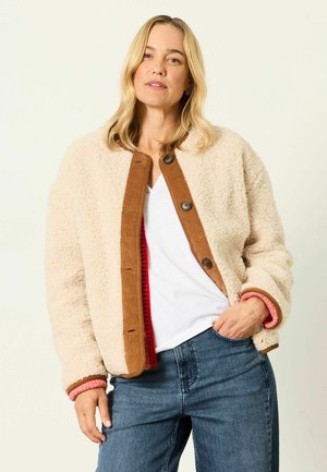 Kvinde iført en beige sherpa jakke med brun kant over en hvid skjorte og blå jeans, stående med den ene hånd i lommen mod en ensfarvet baggrund.